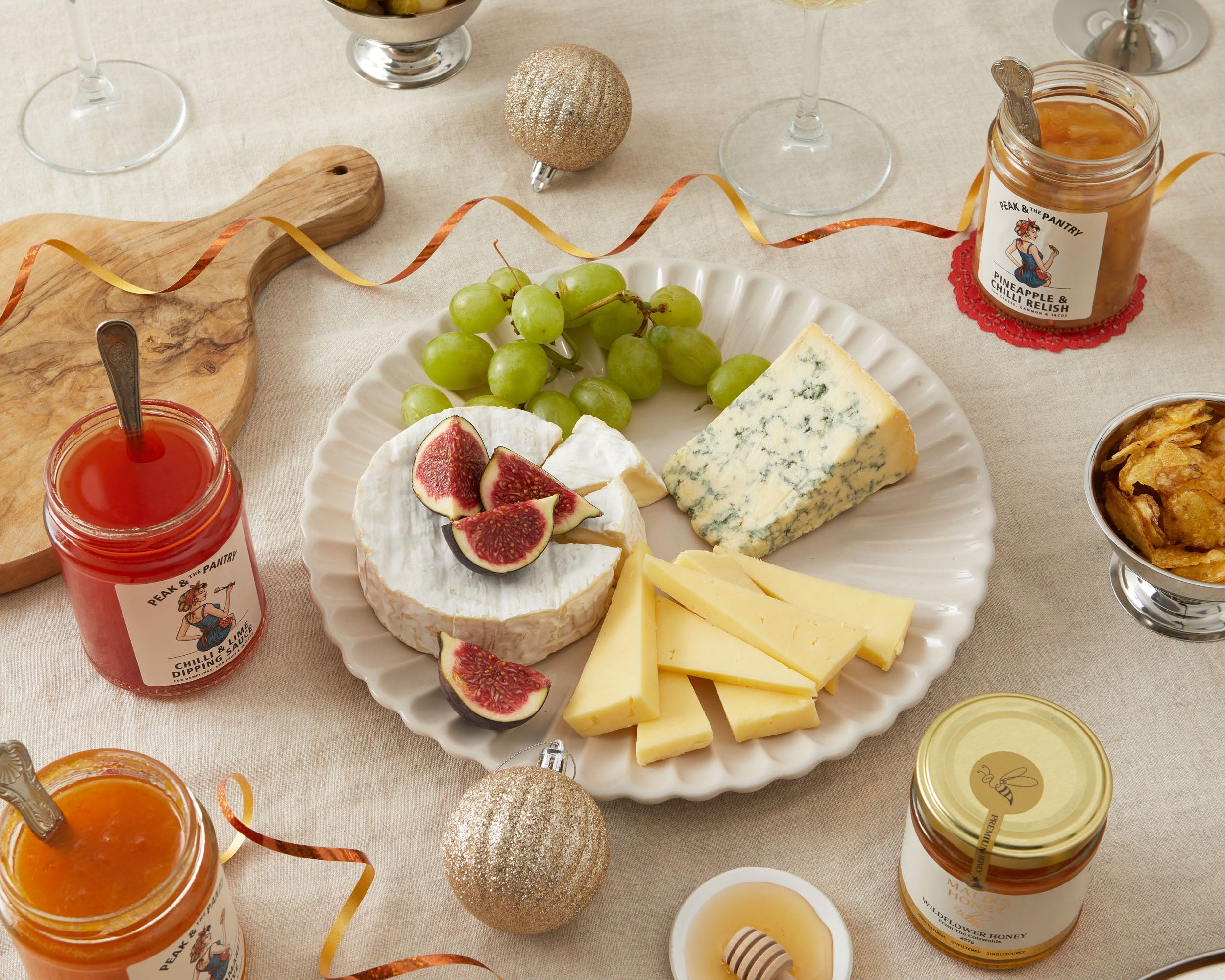 A styled cheese and charcuterie spread on a light beige tablecloth, featuring a white plate with brie, blue cheese, green grapes, figs, and cheese sticks, surrounded by jars of fruit spreads, wine glasses, olives, and decorative ornaments.