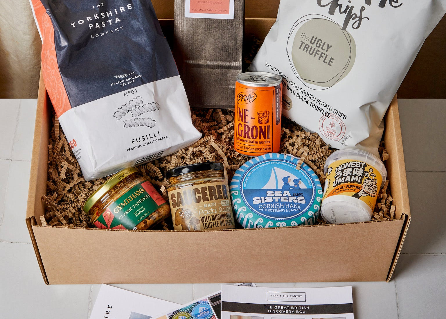 A curated gift box filled with food items, including a bag of Yorkshire Pasta, a bag of Truffle Chips, a can of Mizzoni Crispy Crisps, a jar of cheese, and a round tin, arranged in shredded paper inside a cardboard box. A few printed cards are placed in front on a light surface against a beige curtain backdrop.