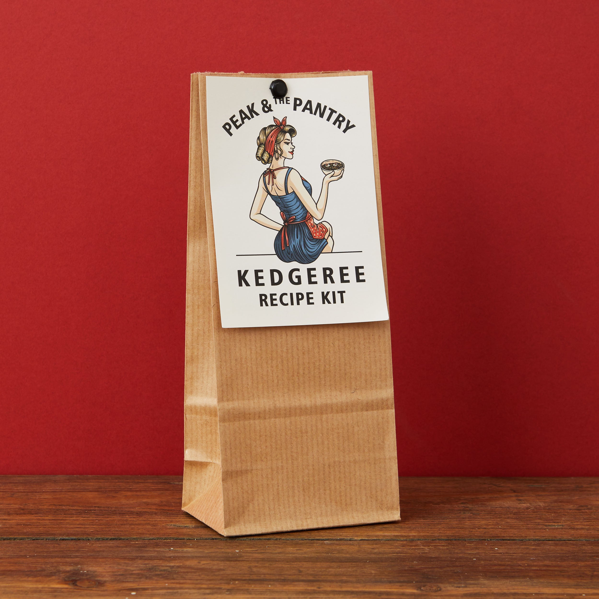 A kraft paper bag package labeled 'Peak & The Pantry Kedgeree Recipe Kit' stands upright on a wooden surface against a solid red background.