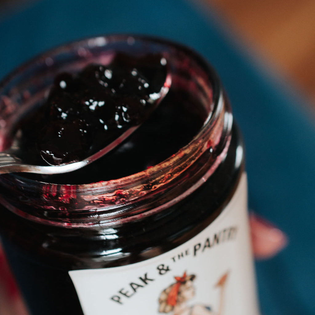 Jar of chuckleberry jam with a spoon, held by a hand, against a blurred background.