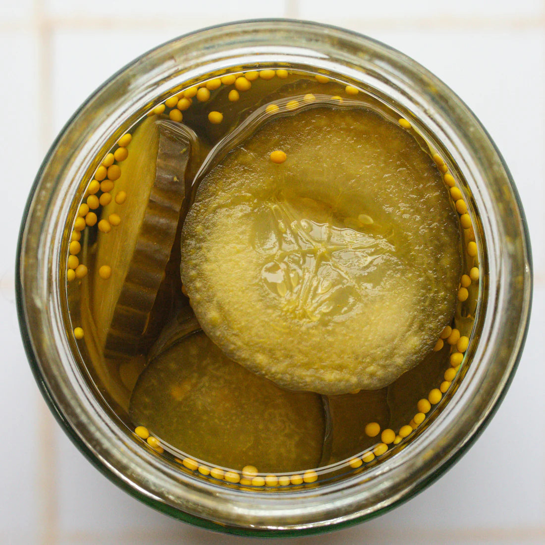 Top-down view of a clear glass jar filled with pickled cucumber slices in a yellow oil, with mustard seeds around the rim, on a white tiled background.