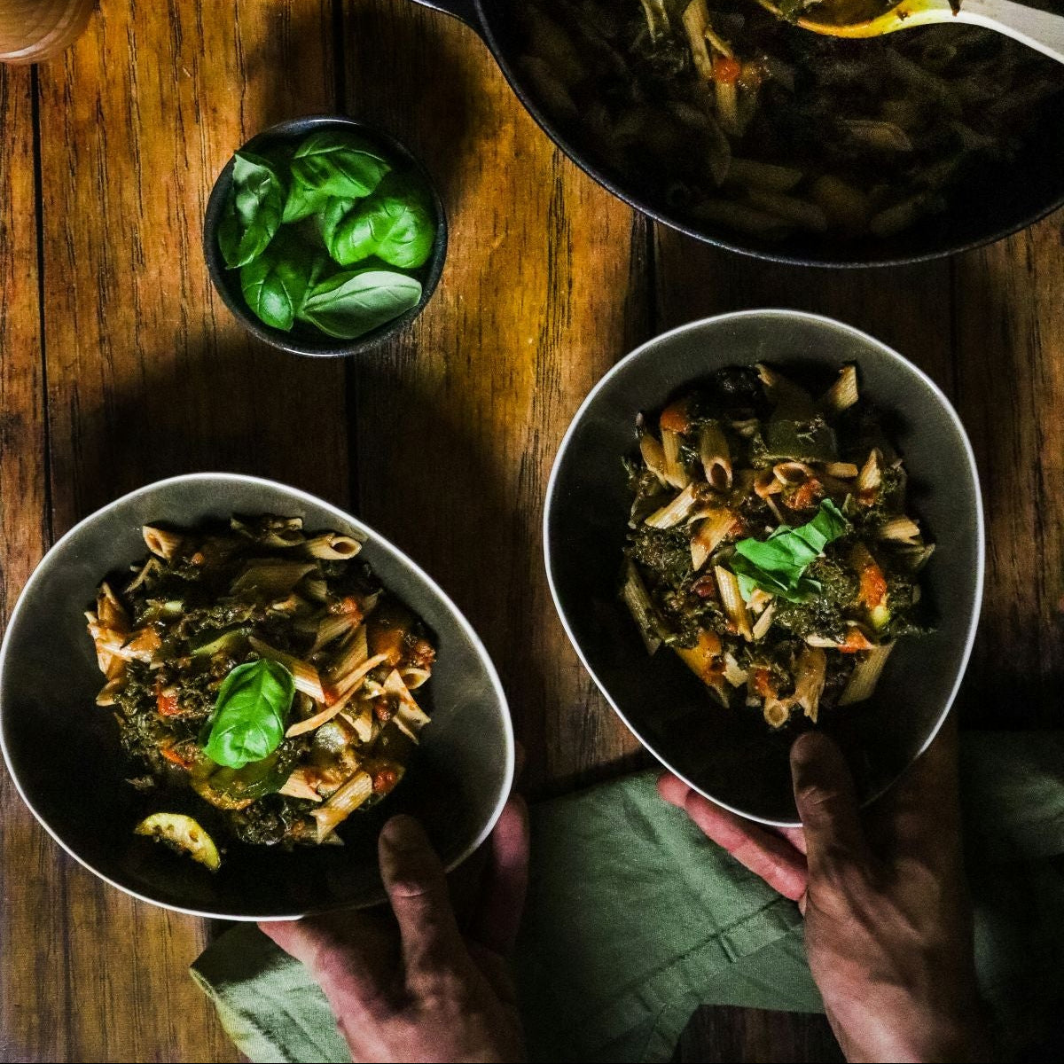 Two bowls of pasta with greens and a pan of pasta on a wooden table.