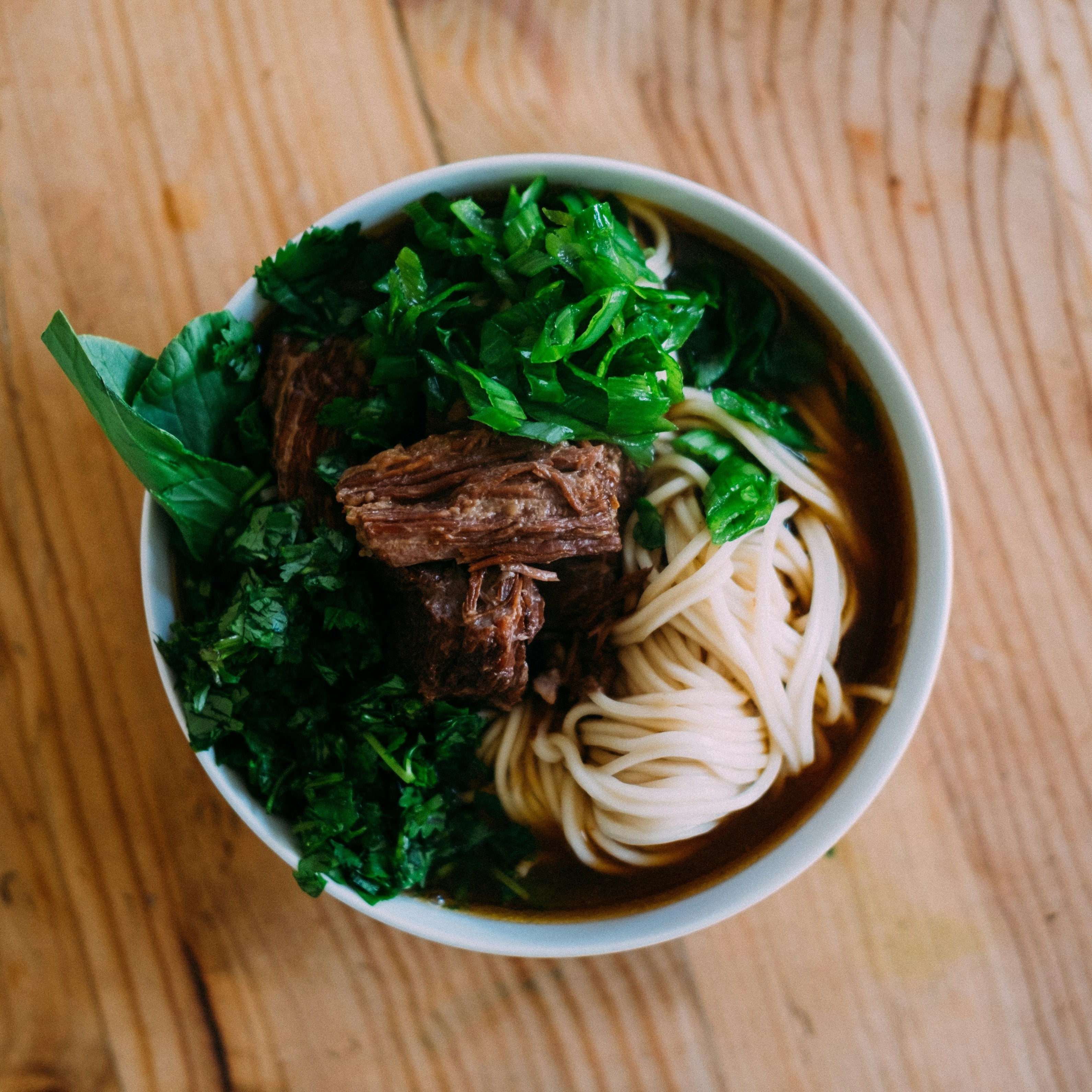 Bowl of noodles with greens and meat on a wooden surface