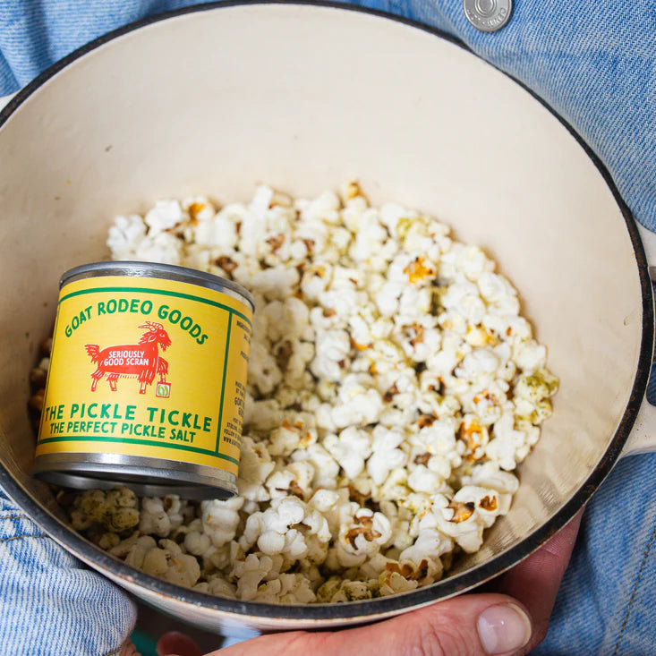 A close-up of a white enamel pot filled with popcorn, with a small yellow tin of pickle salt resting inside. The pot is held by a person wearing a blue denim jacket.