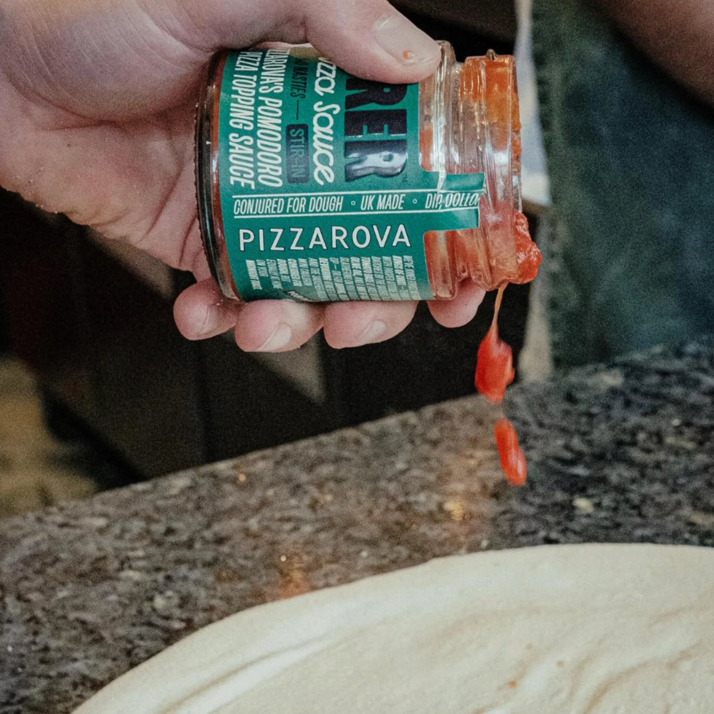 A hand holding an open jar of pizza sauce with sauce dripping out onto a granite countertop next to a round of pizza dough.