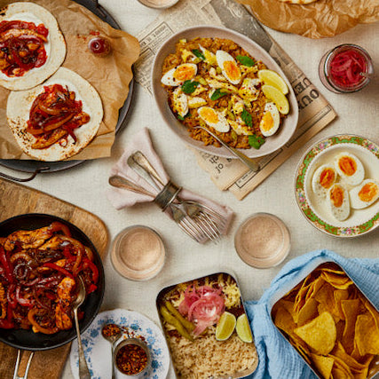 Overhead view of a styled food spread with multiple prepared dishes, tortillas, chips, salsa, drinks, and tableware arranged on a light surface.