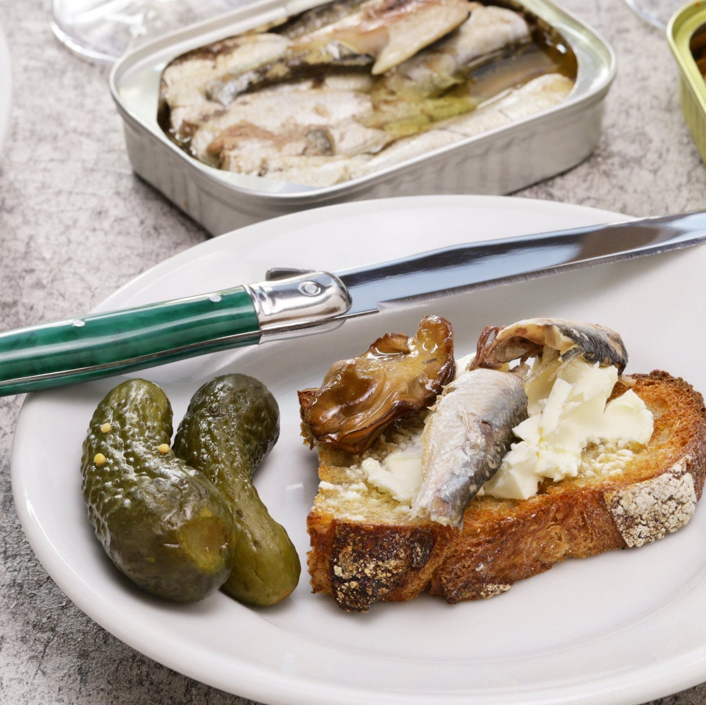 A styled food scene on a gray stone surface showing plated toasted bread, pickles, a knife with a green handle, and two cans of food, including one can of mussels and another of mushrooms, with wine glasses in the background.
